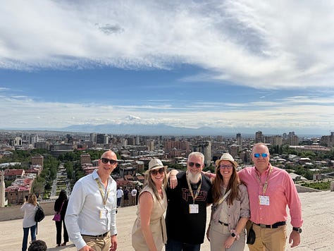 Photos from the Concours Mondial de Bruxelles cultural program in Armenia, including a group dinner at a long candlelit table, a meeting room with the Armenian minister of economy beneath the national flag, a display of Soviet-era wine industry memorabilia and archival photos, aging wine barrels in a cellar, a group photo overlooking Yerevan with Mount Ararat in the distance, illuminated performers at the Garni Temple at night, traditional dancers performing acrobatics, a crowd joining a folk dance circle outdoors, and a large karas amphora covered in visitor signatures." Let me know if you'd like it trimmed or reordered to lead with a particular image.