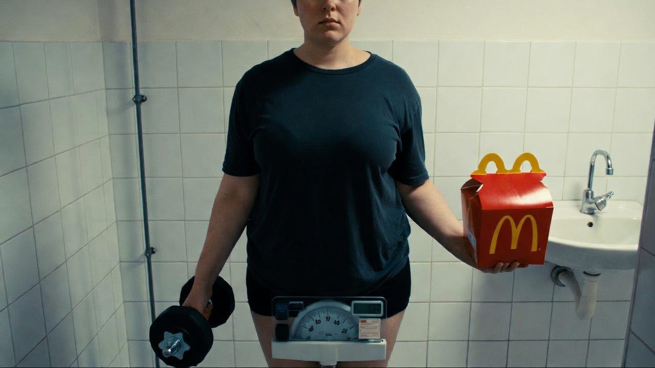 A person on a bathroom scale holds a dumbbell and Happy Meal box in a bathroom setting.