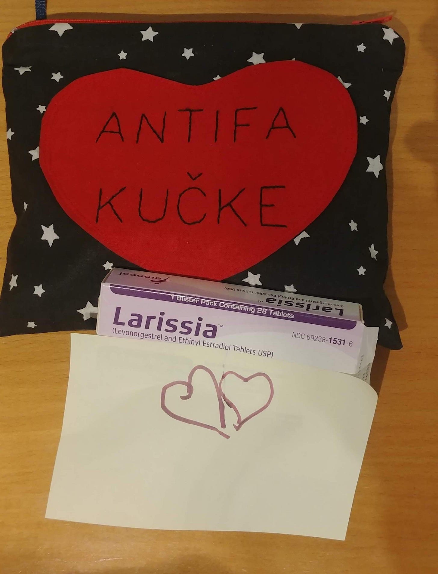 A photo of a table with several objects. At the top is a pencil case made out of black fabric with white stars on it. The front of the pencil case has a heart with "Antifa Kucke" stiched onto it. Below the pencil case is a box with Larissa, a type of birth control. Identifying details on the box are covered with a post-it that has a heart drawn on it.