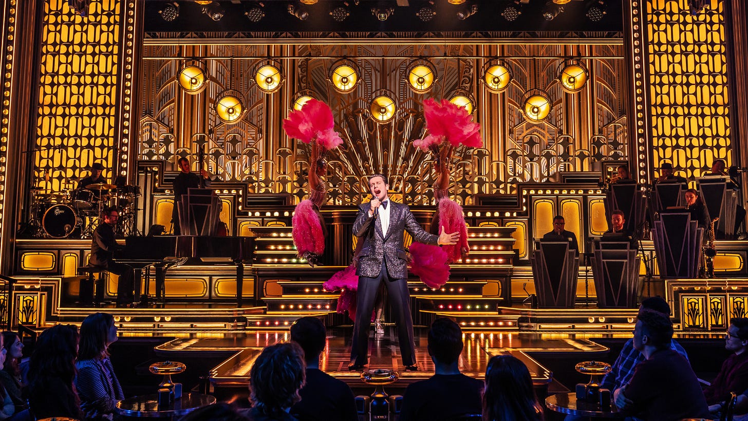 Jonathan Groff sings into a handheld mic as Bobby Darrin. He's flanked by showgirls with pink feathers on a gold nightclub set. Jonathan Groff sings into a handheld mic as Bobby Darrin. He's flanked by showgirls with pink feathers on a gold nightclub set.