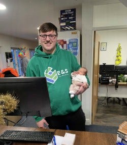 A man in a green hoodie holding a baby stands behind a desk at B&S Building Supplies.  Office supplies and paperwork are visible on the desk and other furniture in the background. 
