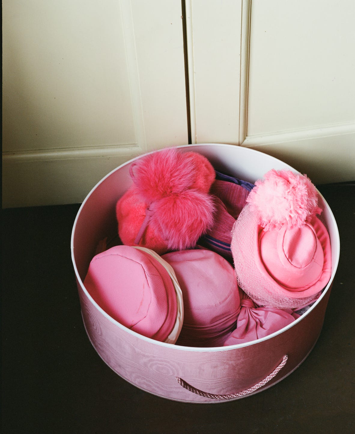 A round box filled with various pink hats featuring fluffy and patterned textures sits on a dark surface against a cream background. Barbara Cartland Claudia Legge Camfield Place 