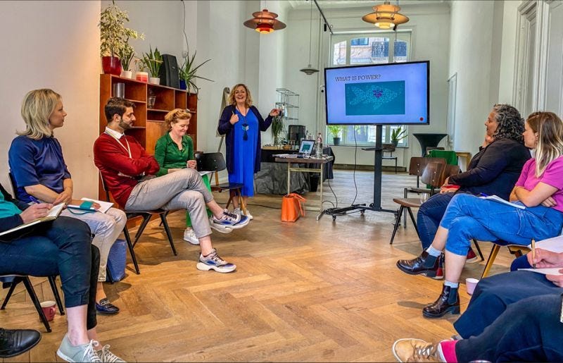 Joanna, a blonde woman with a blue shirt and black cardigan, stands at the front of a presentation and talks to a group of people sitting on chairs in a circle.