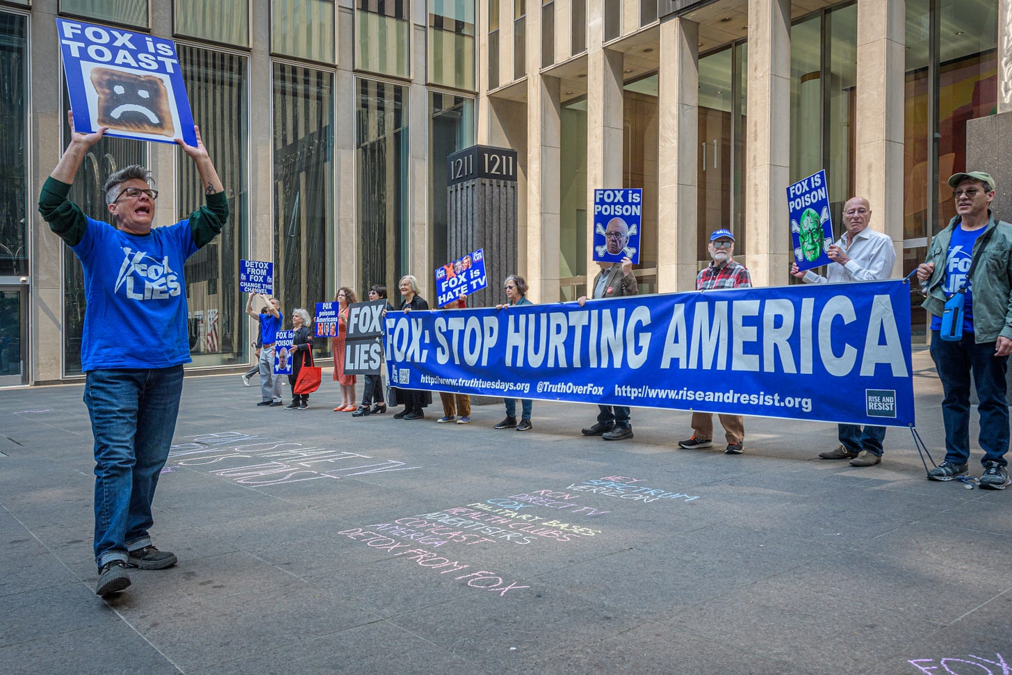 A dozen protestors, several holding a banner that reads "Fox: Stop Hurting America" stand outside a concrete and glass building. In the foreground one protestor holds a sign reading "Fox is Toast" A dozen protestors, several holding a banner that reads "Fox: Stop Hurting America" stand outside a concrete and glass building. In the foreground one protestor holds a sign reading "Fox is Toast"