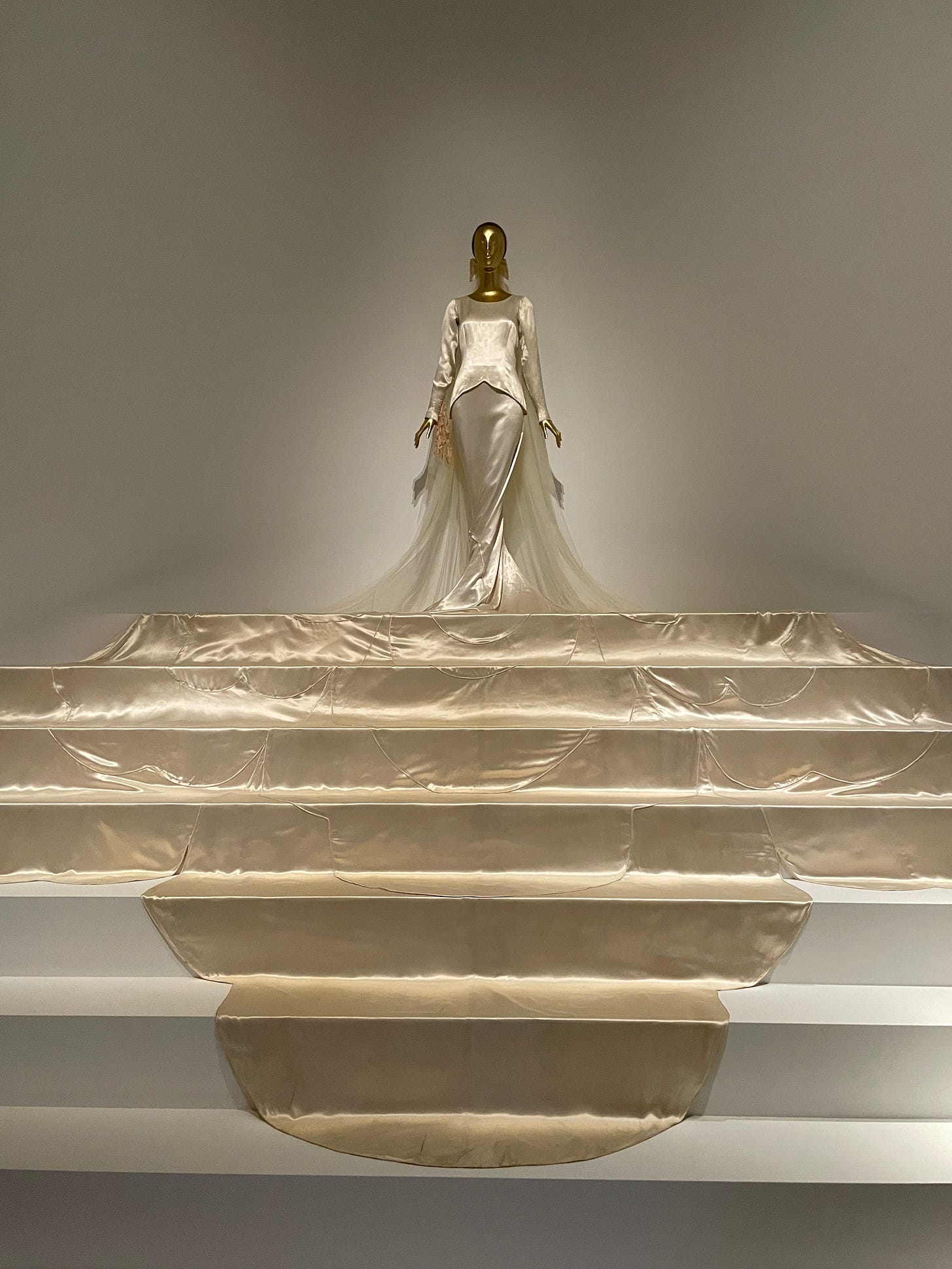 A metallic gold mannequin stands atop a white staircase wearing a cream-colored stain wedding dress with a dramatic train that cascades down multiple steps.