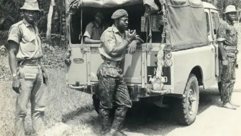 Godwin Alabi-Isama A Nigerian soldier leans against an army truck stopped by the side of a road - thick vegetation seen behind - and speaks into a walkie-talkie. Another combatant can be seen in the back of the vehicle beneath a canvas canopy and two other soldiers are seen standing nearby. It is a black and white archive shot.