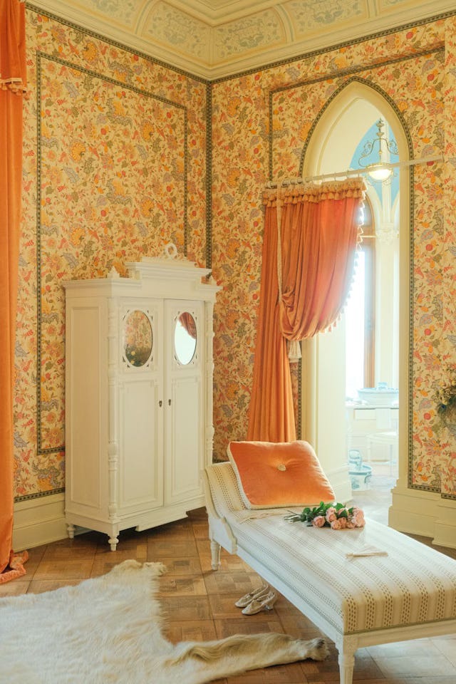 A fairy-tale bed room with peach-colored velvet drapes and a matching pillow on a low, cream colored chaise. An off-white wardrobe with rounded molding details and round mirrors at the top of the double doors stands against the left wall. The wallpaper is yellow and peach ferns with dark grey leaves on an off-white background and has thin greenish grey molding details. The ceiling is arched with painted moldings. A single doorway beyond the curtain leads into another, very bright room.