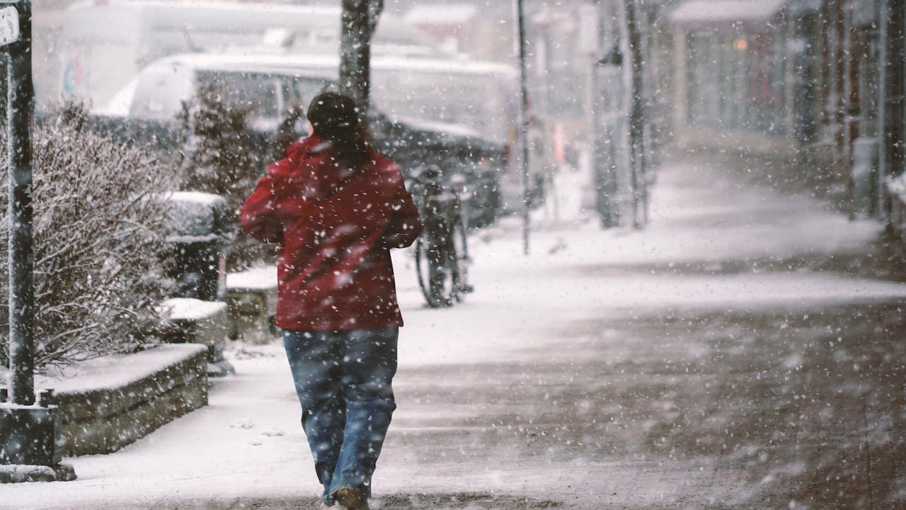 A person with long hair and a red jacket and jeans walks through the snow on a sidewalk. A person with long hair and a red jacket and jeans walks through the snow on a sidewalk.