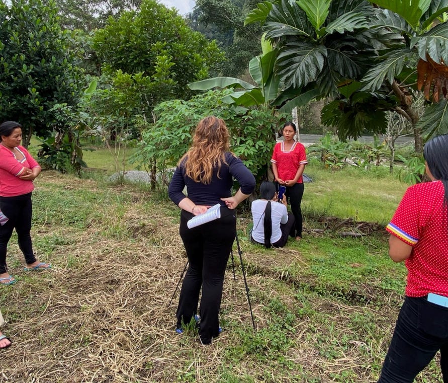 Margot, a blonde European woman in black jeans, stands with her back to the camera while she films a member of the Yuturi Warmi speaking. two other women stand watching