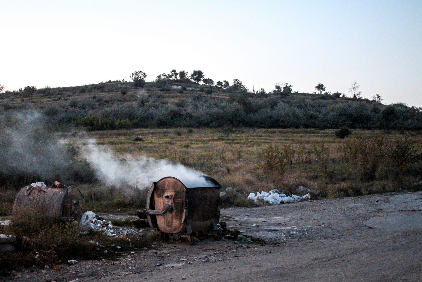 Smoke rises from a rusted skip, litter is strewn around it.