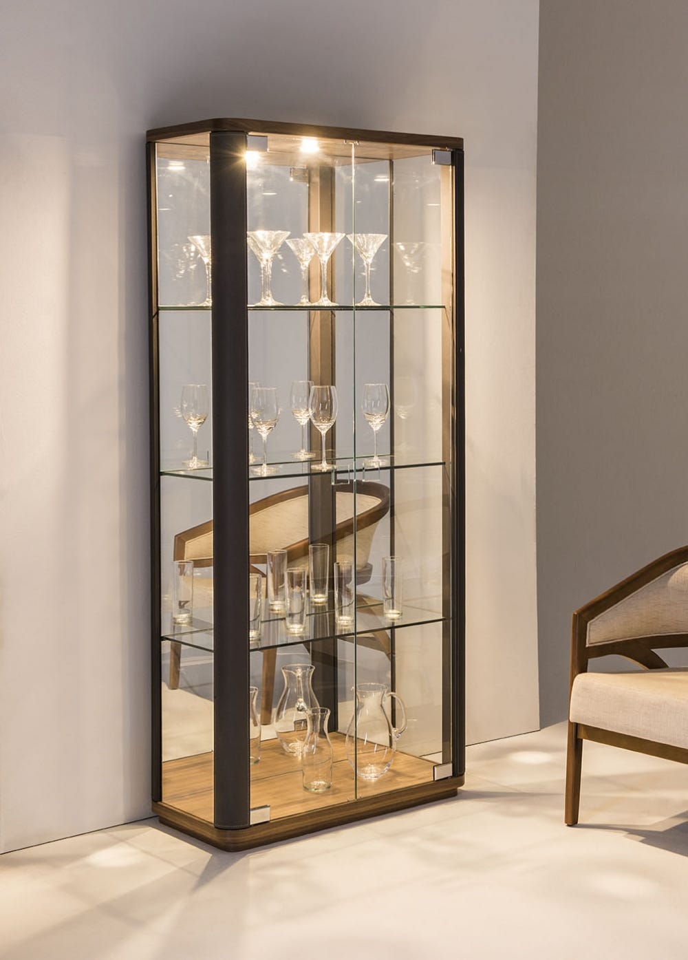 na parede de uma sala iluminada, detronte a uma poltrona, uma cristaleira de madeira toda de vidro, com iluminação interna e diversos copos, taças e jarros de cristal 