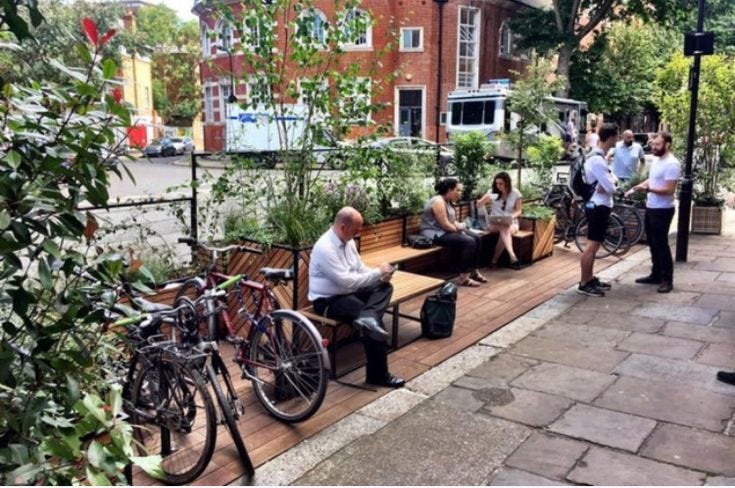 This may contain: people are sitting on benches near bicycles and plants in the city park, while one man is using his cell phone