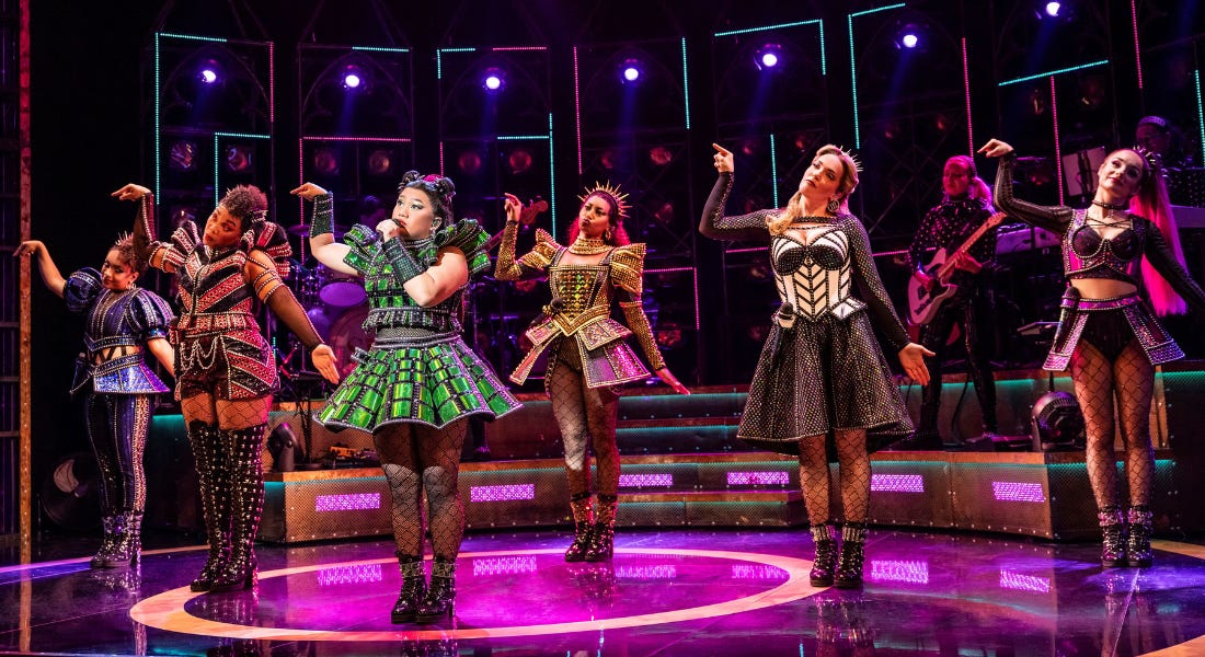 A group of six singers on stage wearing colourful dresses and fish nets, with green and purple lights around them