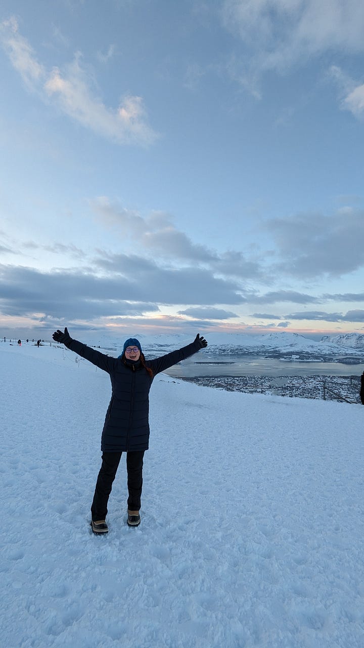 A woman stands with her arms upstretched, delighted, atop Fjellheisen mountain in the snow. Also, Tromso harbour, boats clinking on the still, glassy water of the fjord.