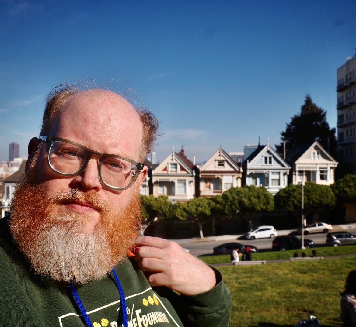 The author standing in a park, head tiltled slighting, staring into the lens, a row of houses in the background. The author standing in a park, head tiltled slighting, staring into the lens, a row of houses in the background.