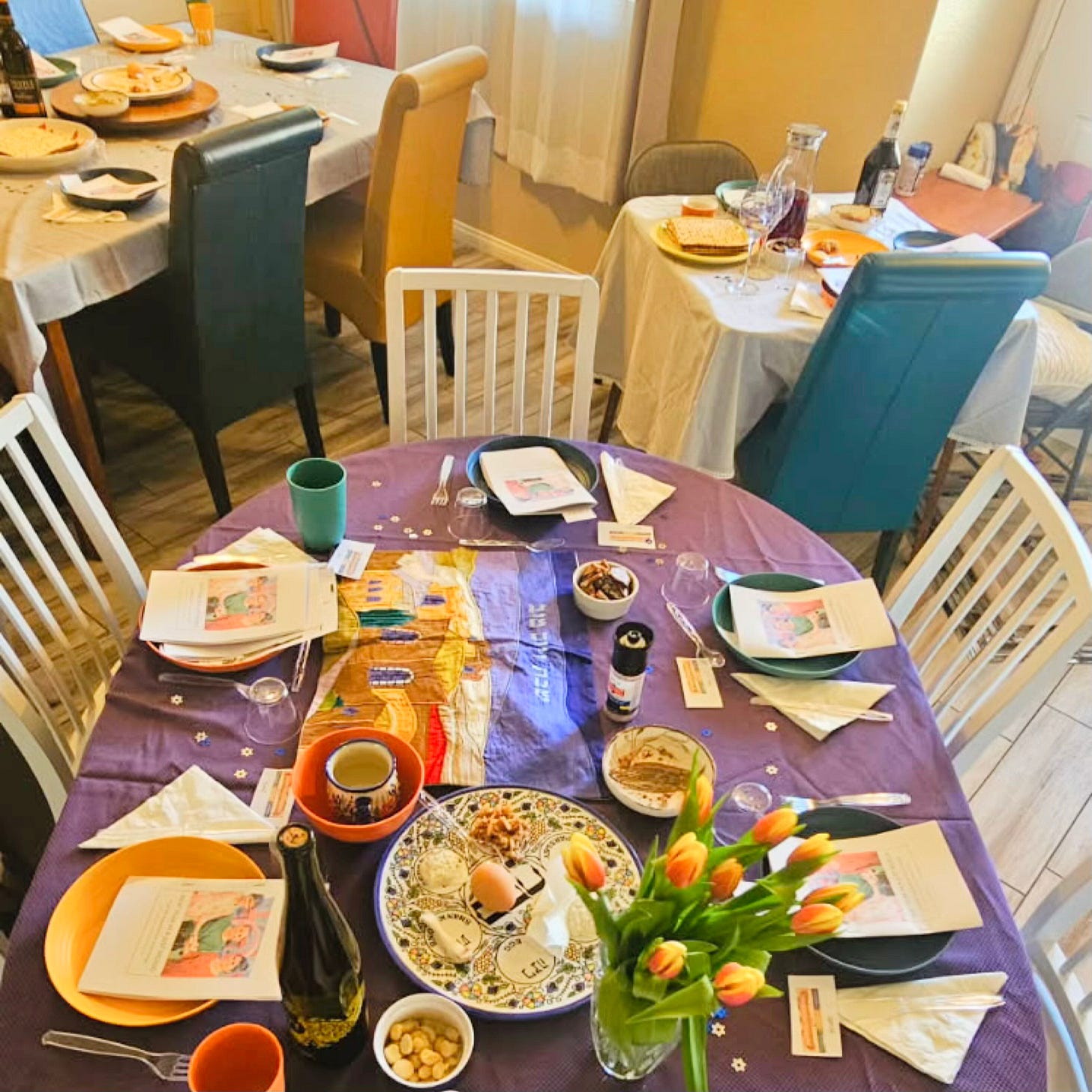 A multi-table Passover Seder setup in a Las Vegas home featuring Haggadah booklets, ceremonial foods, wine, and shared seating arrangements designed for a diverse group of women and their families experiencing Jewish traditions together.