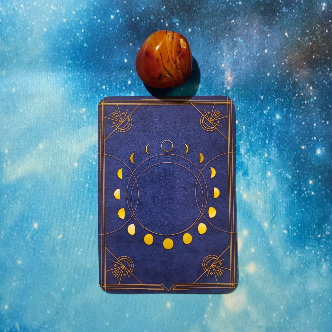 New Moon Magic: Your August 2025 Pick-a-Card Tarot Reading, pile 2 — silk agate (orange and brown) stone and an oracle, face down on a starry blue background
