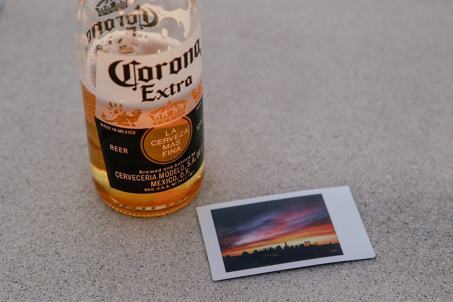A close-up shot of a Corona Extra beer bottle sitting next to a small, instant-style photograph. The photograph itself shows a colorful sunset over a city skyline. Both items are resting on a grey, textured surface.