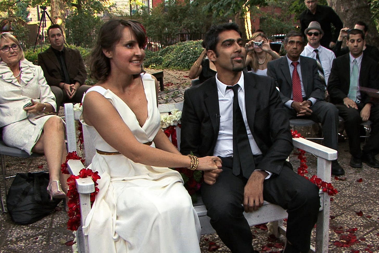 a white woman in a wedding dress and a man of color in a suit sitting on a bench; guests behind them. they look happy and are holding hands