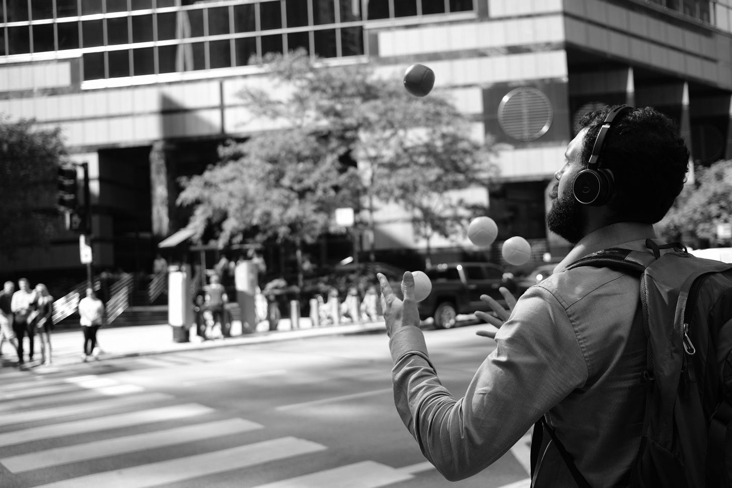 gray scale photo of a man with bag and headphones jugling balls in the air while crossing the street