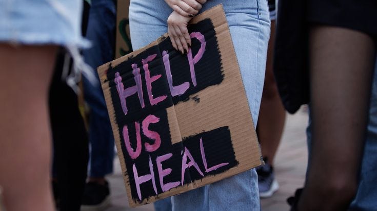 This may contain: a person holding a cardboard sign that says help us heal with pink writing on it