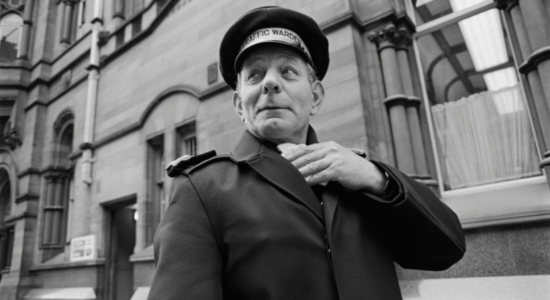 A black and white photo of a traffic warden with a brick building behind him
