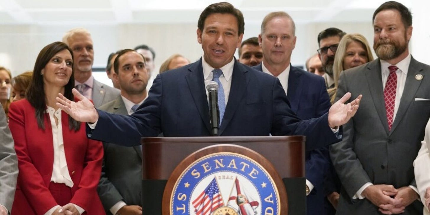 Gubernur Ron DeSantis di depan para wartawan dengan dikelilingi oleh para staff.