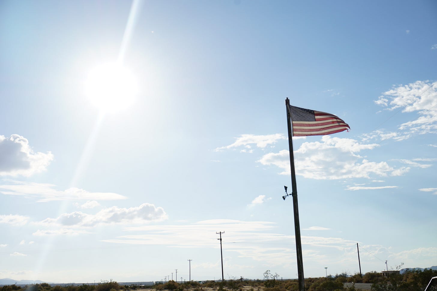 American flag on a tall pole waving in the wind beneath a bright desert sky, with power lines and scattered buildings in the distance.