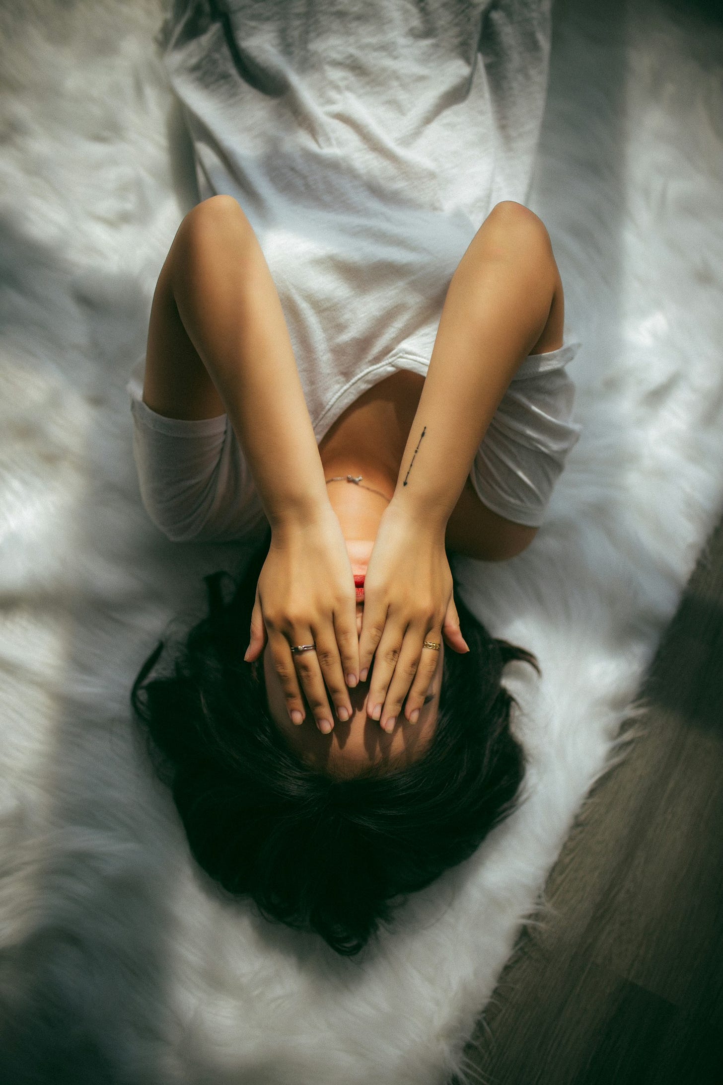 A woman lying upside down on a white comforter, wearing a white v-next shirt, hands over her face.