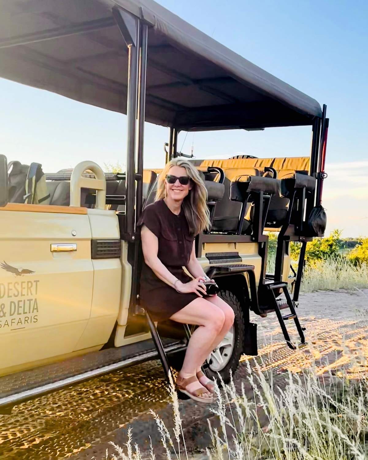 A woman sits on the steps of a safari jeep in Africa, She is wearing a short brown dress and holding a camera.