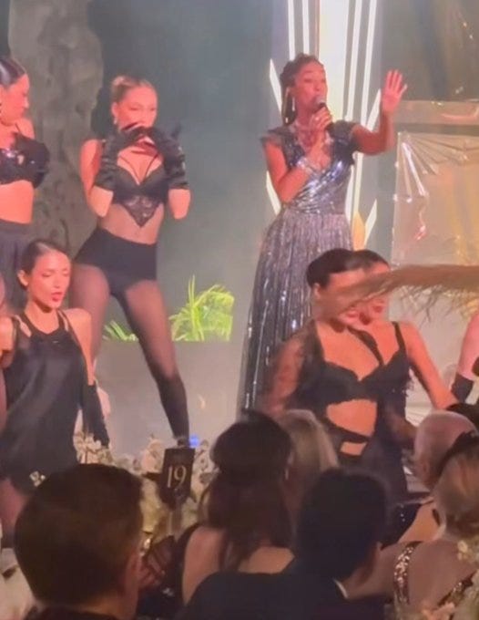 Group of women in revealing performance outfits including black lace tops, sheer dresses, and gloves pose and gesture on a lit stage surrounded by palm plants and metallic pillars. Central figure in sparkling gown raises hand. Number 19 visible on foreground stand. Diverse ethnicities among performers with long hair and makeup.