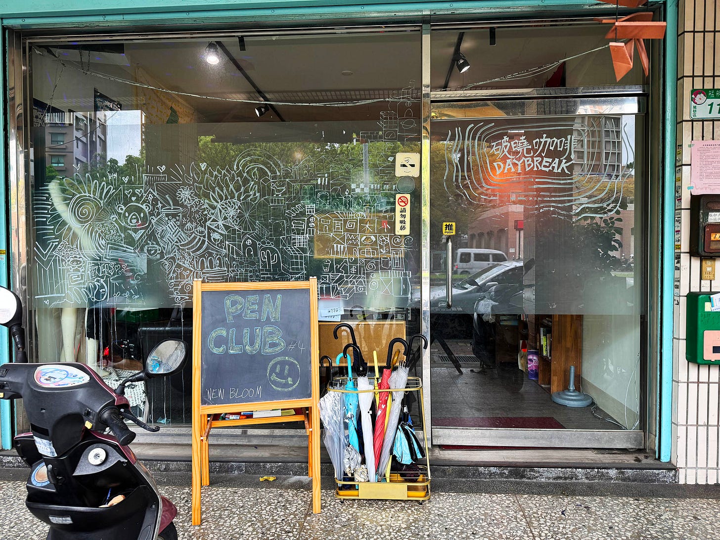 Horizontal photo of the Daybreak space storefront in Wanhua, with hand-drawn art on the glass front, and an A-frame chalk board sign that says Pen Club.