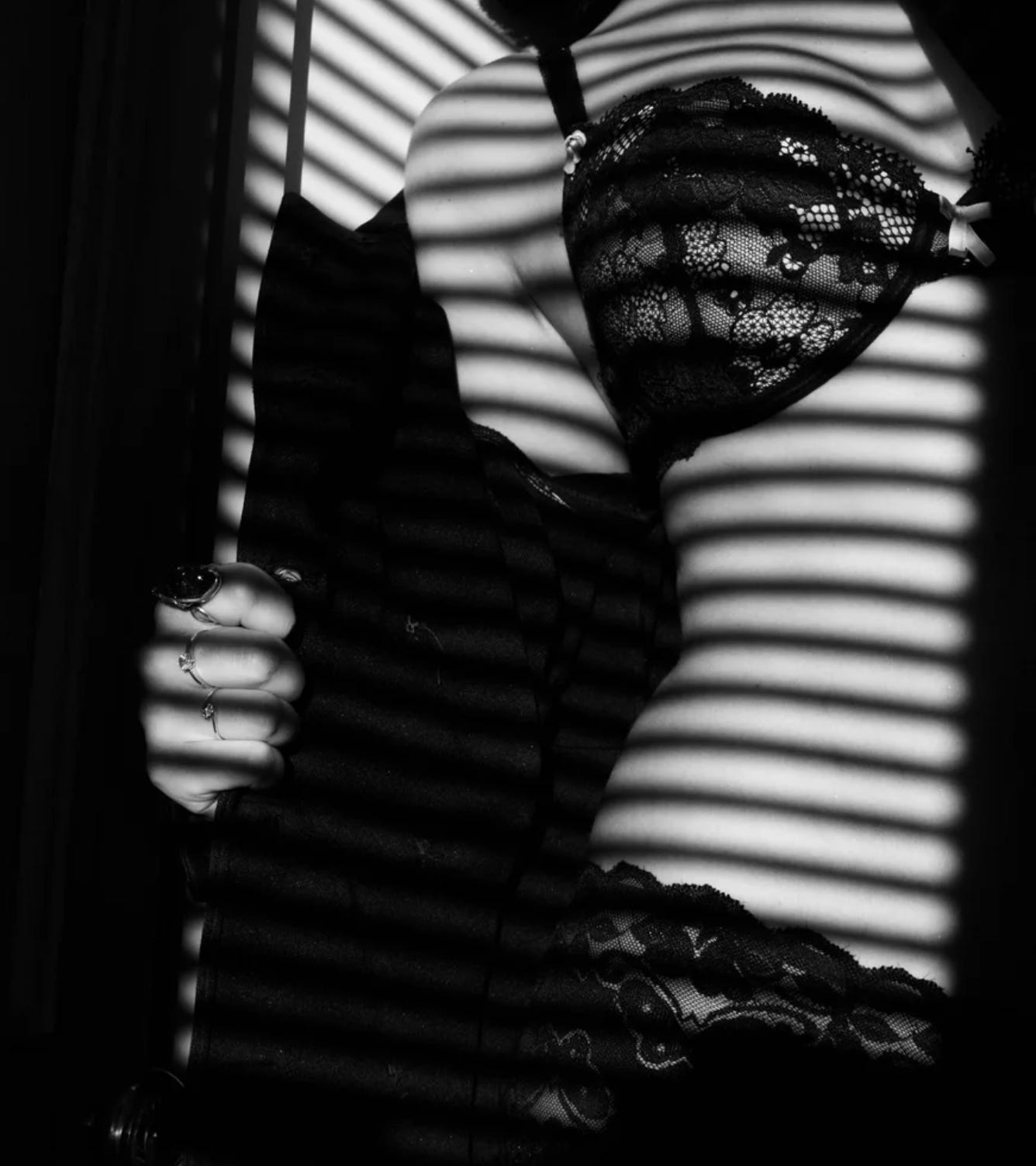 A woman in lingerie stands in a window with the reflection of the blinds on her body