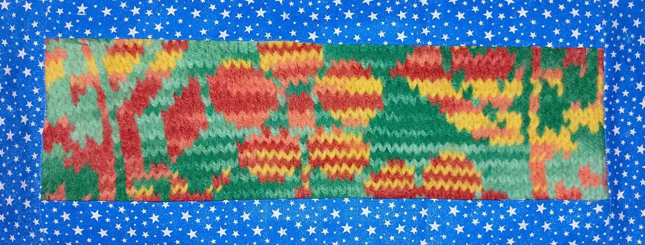 A imagem mostra uma pequena tapeçaria em malha de tricô com um design abstrato e colorido.  A peça apresenta padrões que lembram flores estilizadas e formas representando folhagem. As cores vibrantes incluem tons de verde, vermelho, amarelo e laranja. A borda externa do tecido é de um papel azul escuro com pequenas estrelas brancas.