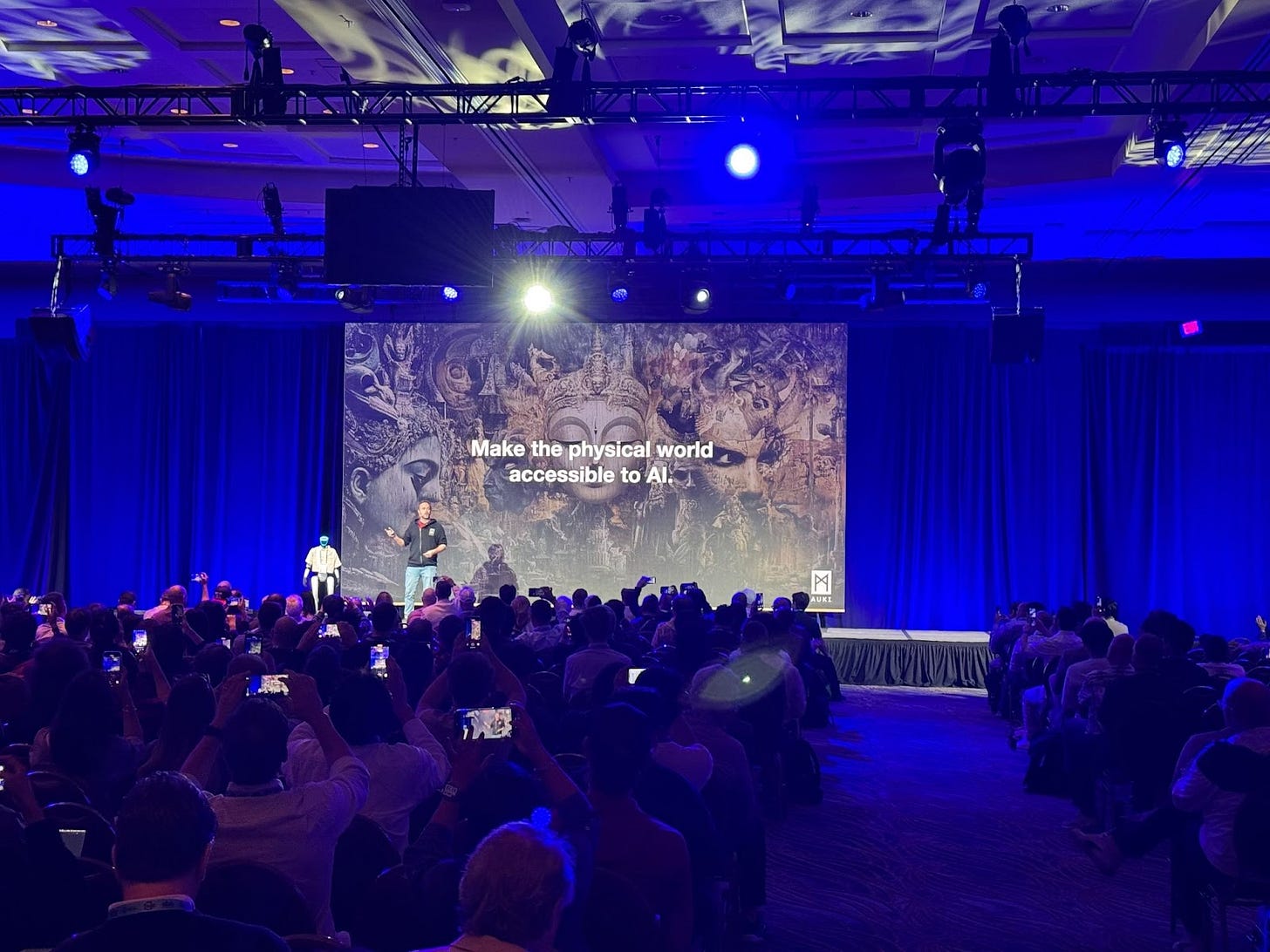 A speaker stands on stage at a packed conference with a large screen behind them displaying the words, "Make the physical world accessible to AI." The audience is seated in rows, many holding up phones to capture the moment. The room is dimly lit with blue and purple stage lighting, and the screen features an artistic background of intricate sculptures and faces. A speaker stands on stage at a packed conference with a large screen behind them displaying the words, "Make the physical world accessible to AI." The audience is seated in rows, many holding up phones to capture the moment. The room is dimly lit with blue and purple stage lighting, and the screen features an artistic background of intricate sculptures and faces.