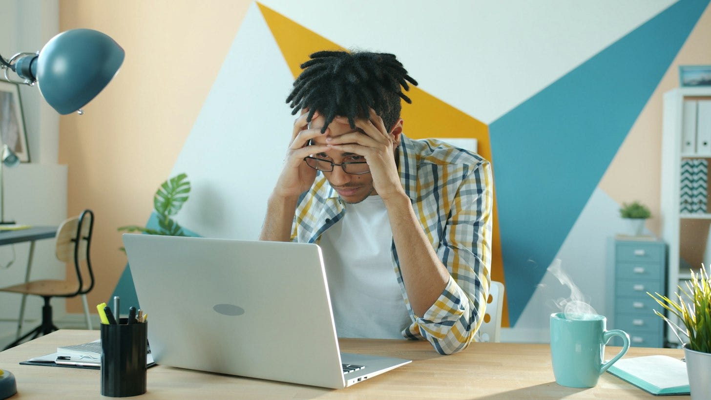 A man with his hands pressed to his head sits at a desk in front of an open laptop, looking down at the screen. A man with his hands pressed to his head sits at a desk in front of an open laptop, looking down at the screen.