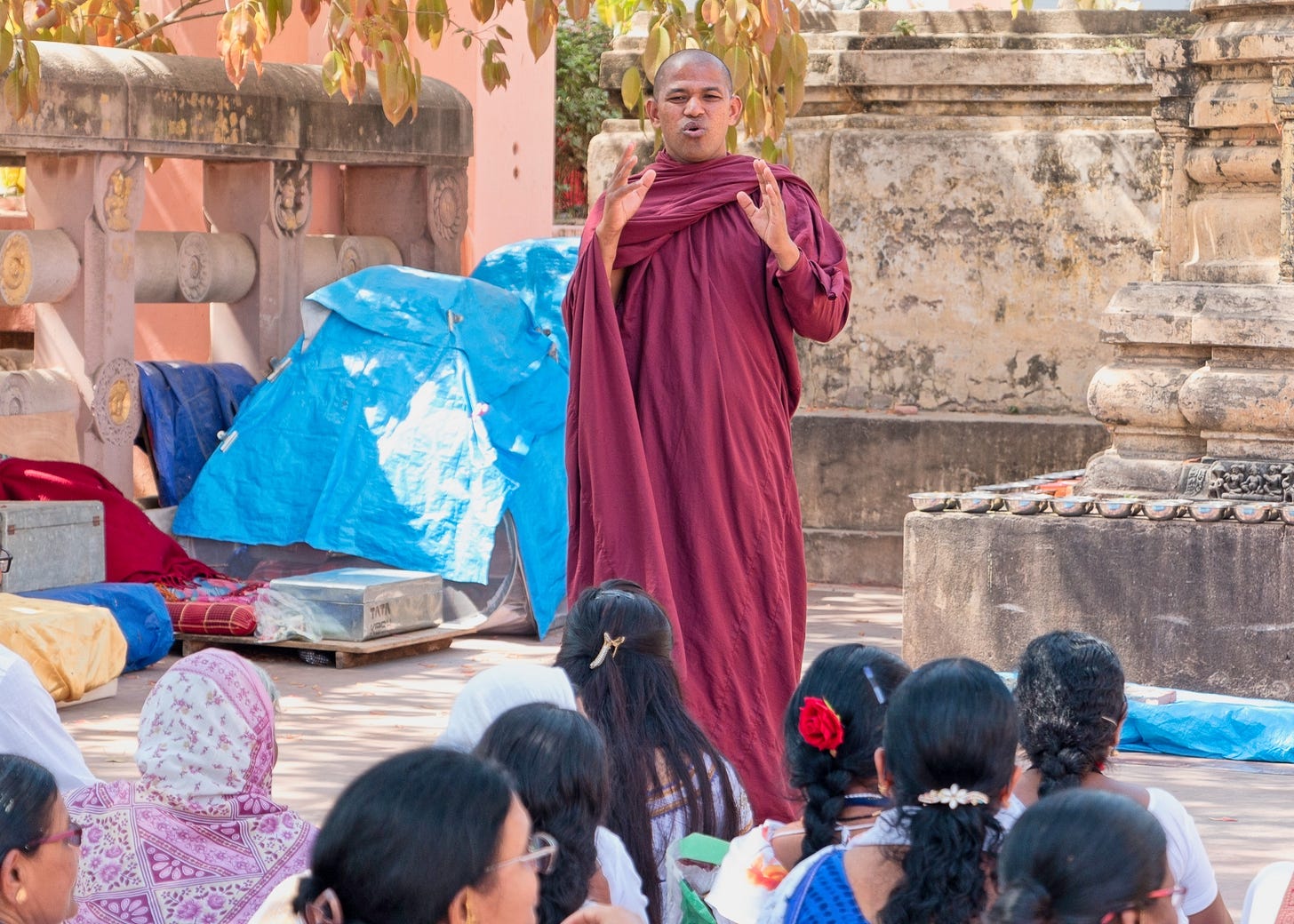 Man in maroon robe addresses seated visitors outside stone temple Man in maroon robe addresses seated visitors outside stone temple