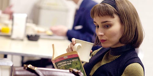 cena do filme Carol: a personagem Therese de cabelos castanhos lê o livro Retrato de uma artista quando jovem (em inglês)