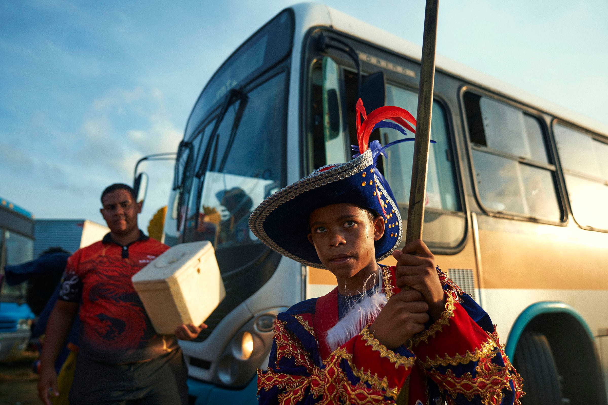 A young person in a colorful, feathered costume and hat holds a staff in front of a bus, while another person in casual clothing stands nearby holding a white box. The scene is lit by evening sunlight.