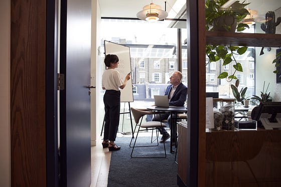 A woman at a whiteboard talking with a man in a business suit sitting down. A woman at a whiteboard talking with a man in a business suit sitting down.