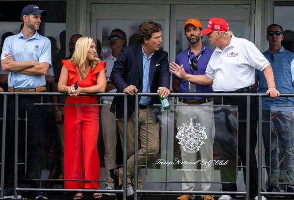 Tucker Carlson and Donald Trump stand and talk to one another on a balcony with a few people beside and behind them.