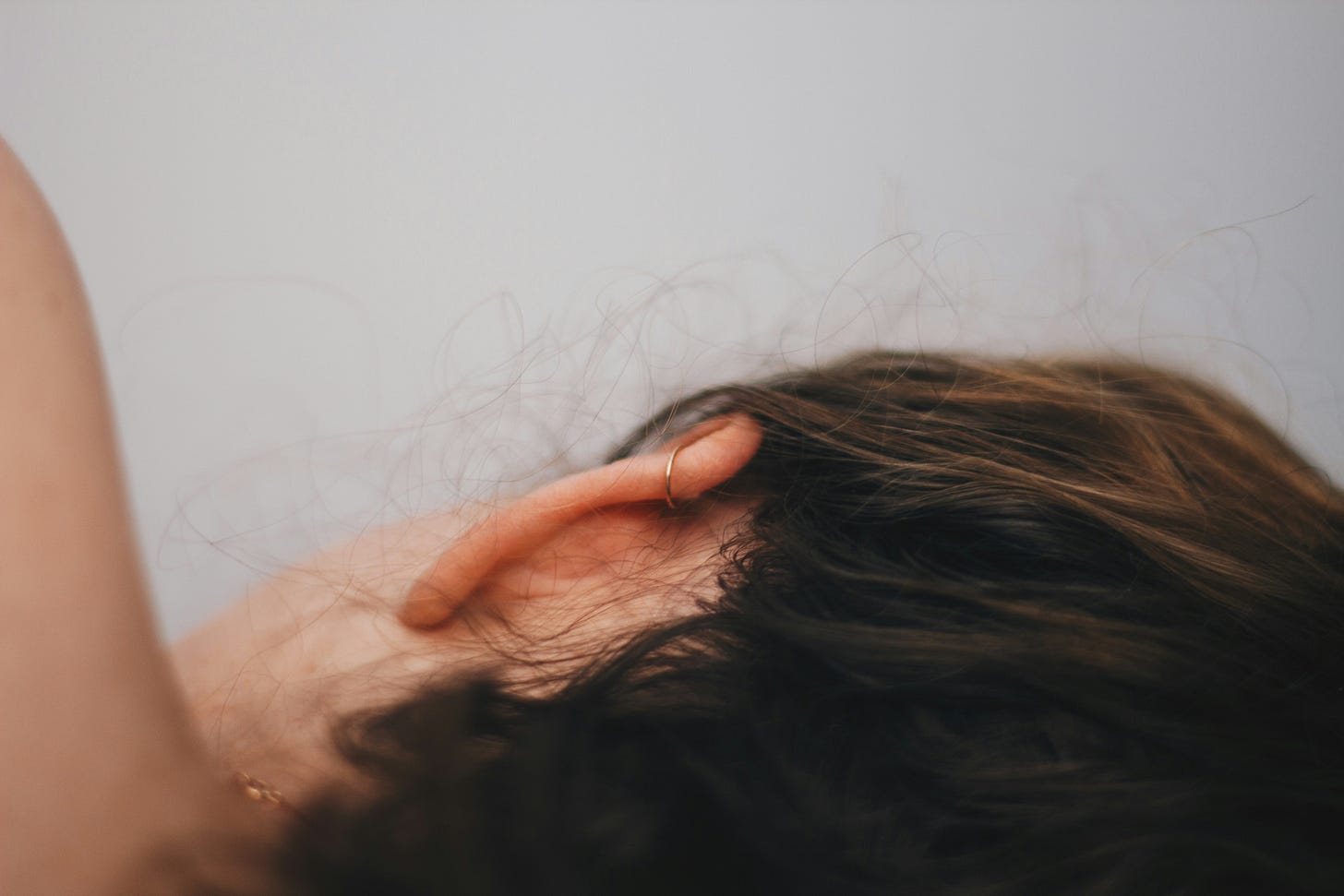 Close-up of the back of a white person with brunette hair’s head. They are lying on their right side. They are wearing a gold cartilage hoop in their ear and a necklace.