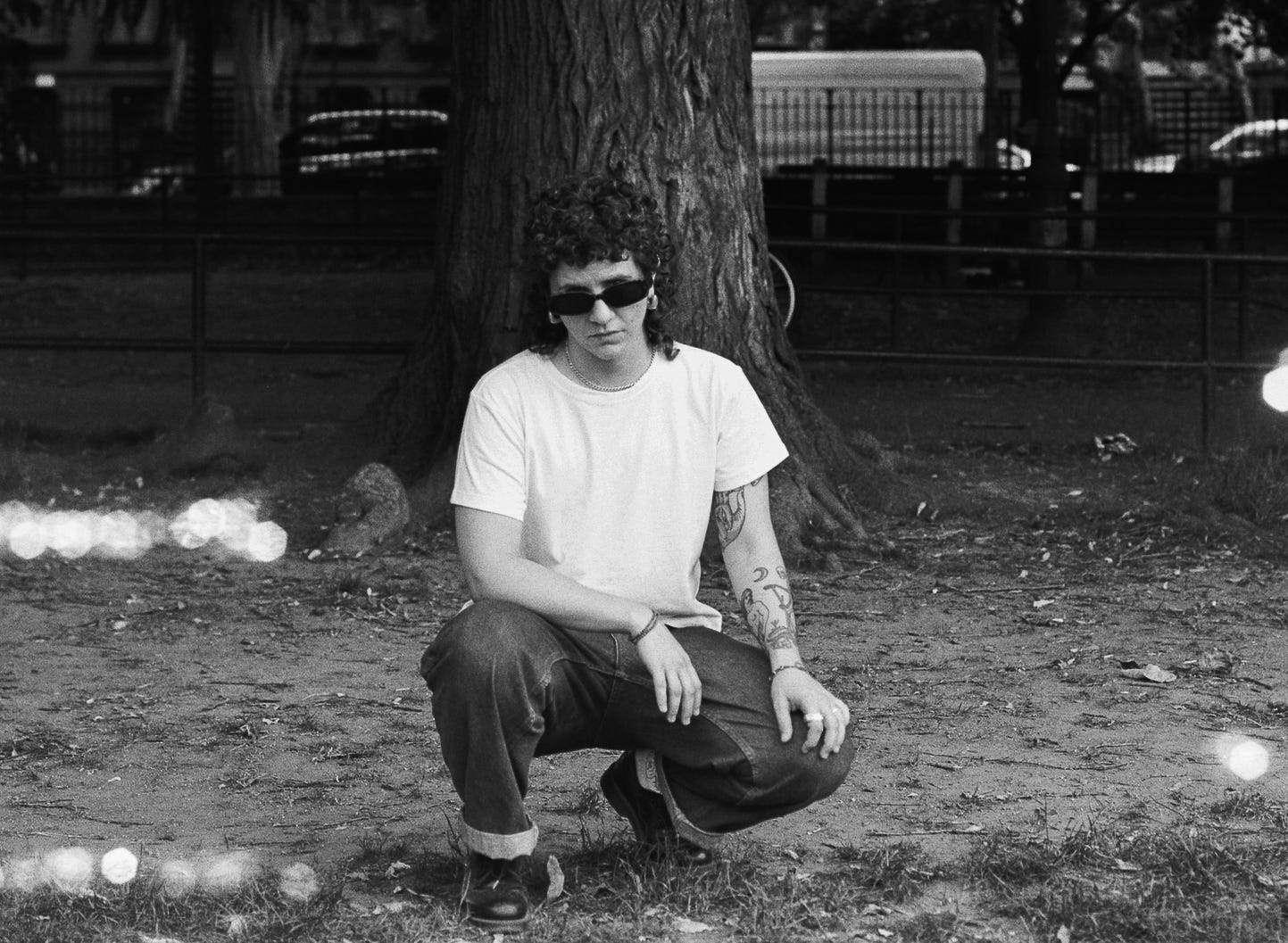 Lou Lauren crouches at the base of a tree in a white tee and loose denim, gazing forward in dark sunglasses. The energy is soft and grounded, with dappled light marks streaking the frame giving the image a raw, almost spectral analog feel. 
