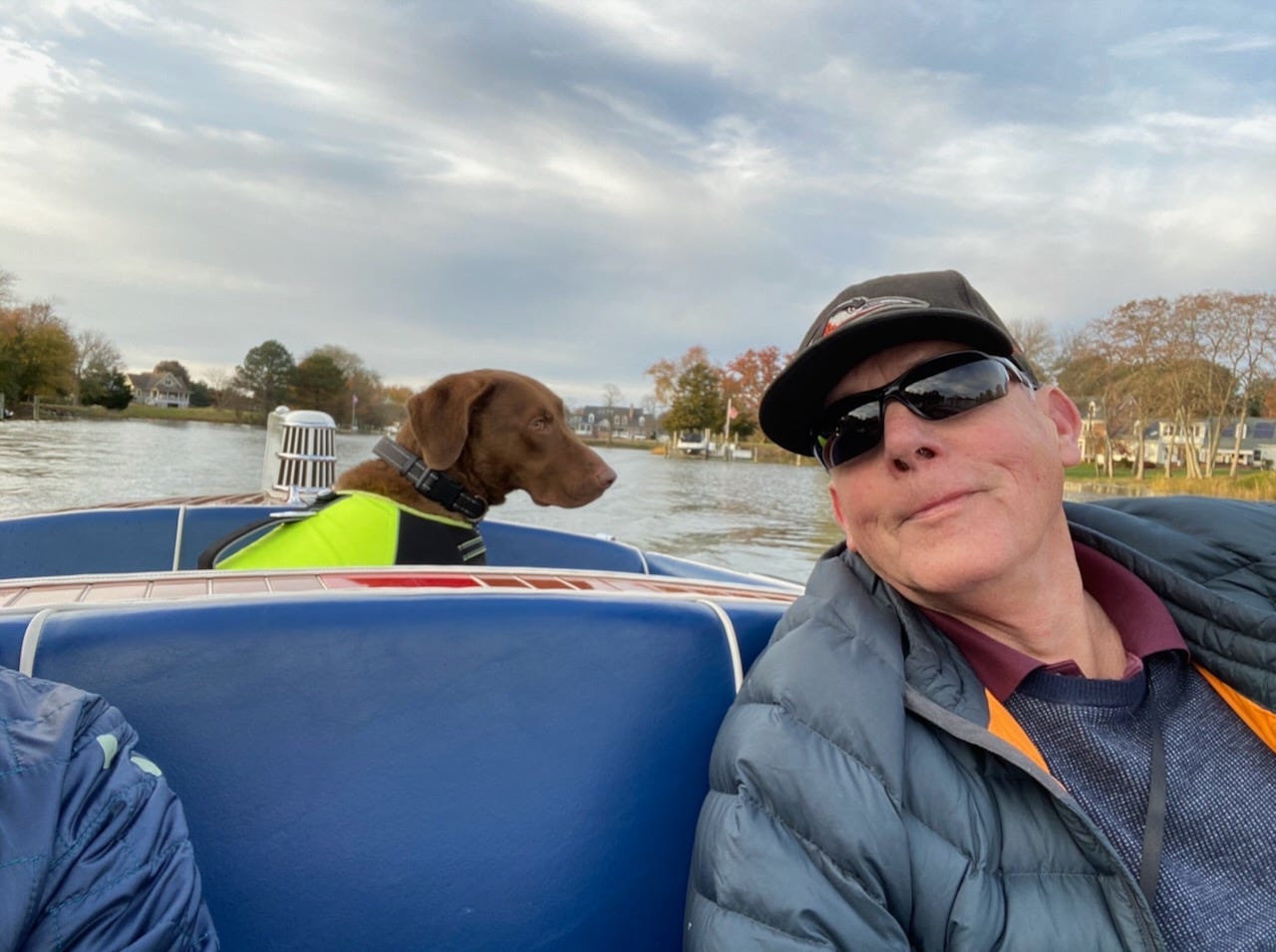 A brown dog in a yellow life jacket sits in the stern of an antique wooden power boat on calm water, with houses and autumn trees visible along the shoreline.