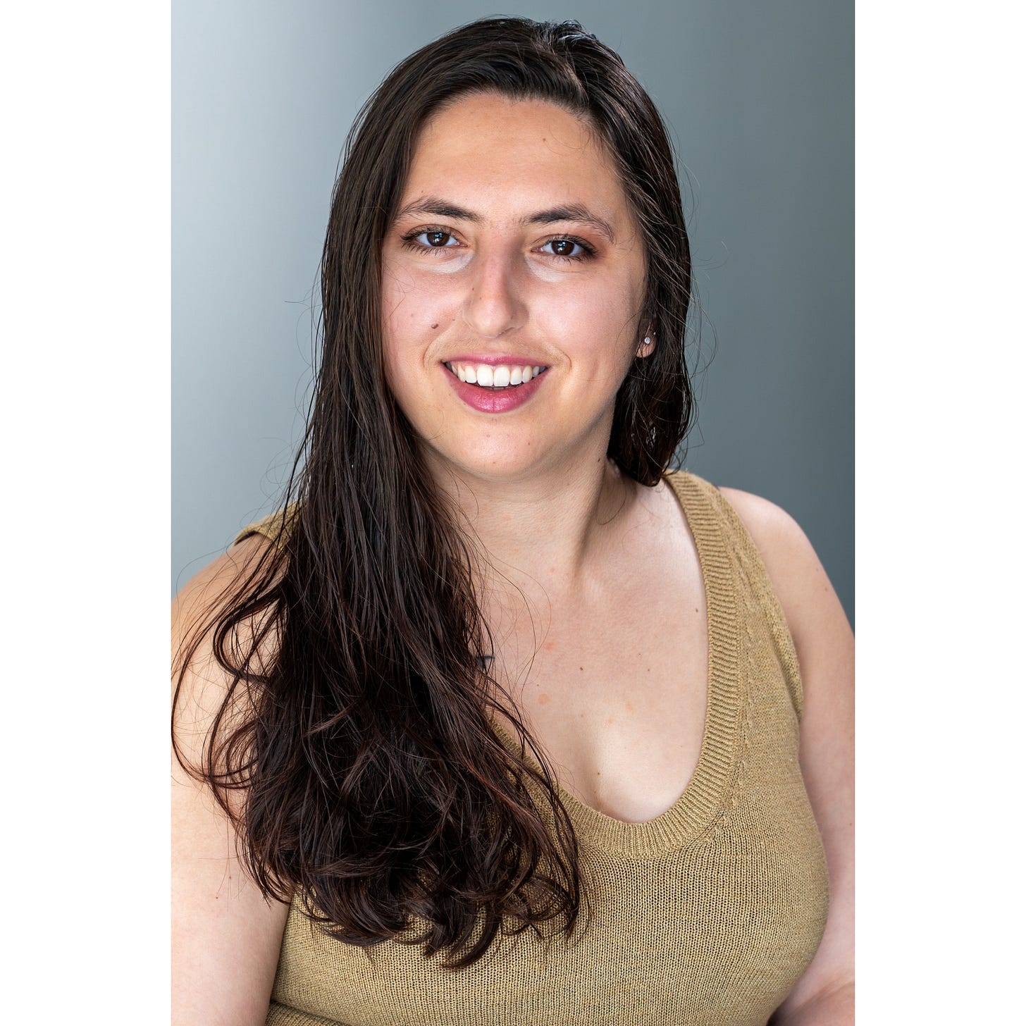 Headshot of Sonya Rio-Glick: a lightskinned woman with long brown hair smiles at the camera. She is wearing a tan sleeveless top against a gray background.
