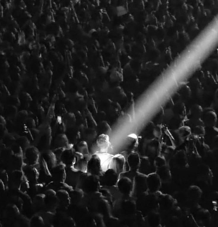 This may contain: a large group of people at a concert with one person on stage and the light shining down