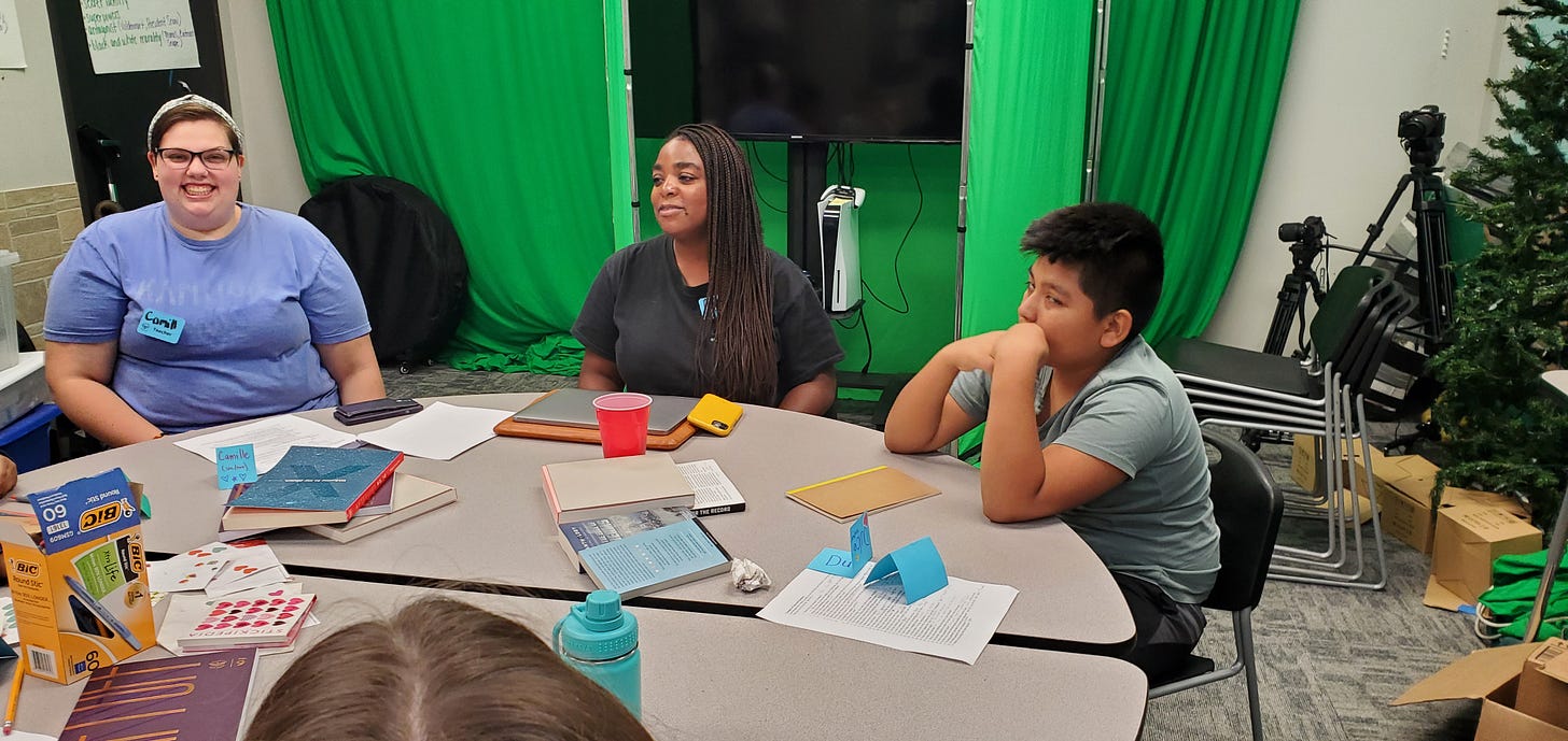 ABC Romantasy Camp instructors and students gather around a gray table in a classroom to discuss the writing activity for camp.                                                        Los instructores y estudiantes en el campamento de fantasia romántico se reúnen alrededor de una mesa gris en un salón de clases para discutir la actividad de escritura para el campamento.