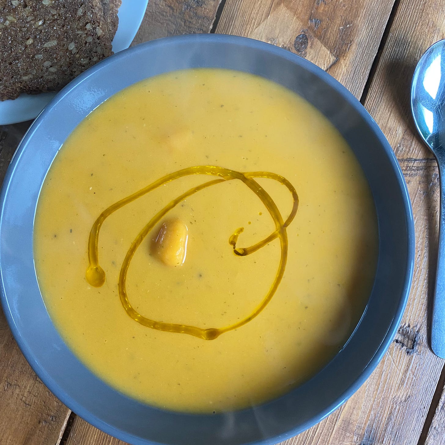 Bowl of Sweet potato, coconut and chilli soup on a wooden table, with a sooon placed to the right and some rye bread in the to left corner