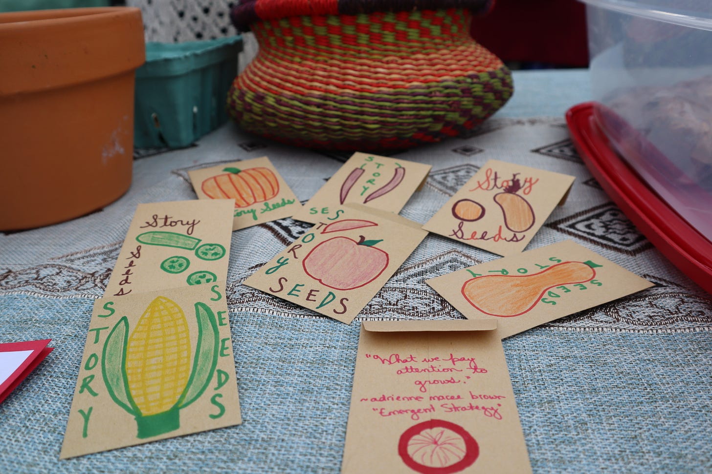 Hand-decorated brown paper seed packets with colorful drawings of produce, the words "Story Seeds," and a quote by adrienne maree brown: "What we pay attention to grows." 