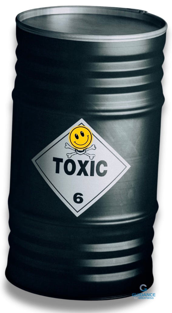 A dark gray metal barrel with horizontal ridged bands around its middle section. The barrel displays a white diamond-shaped hazard label marked "TOXIC" in bold black letters with the number "6" below. Above the text is a bright yellow smiley face emoji with crossbones beneath it, creating an ironic juxtaposition between the cheerful symbol and the toxic warning message.